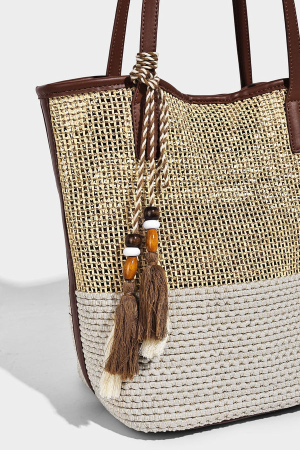 Woven straw-look tote bag in tan and cream with dark brown leather-look trim, long flat shoulder straps, and tassel bead