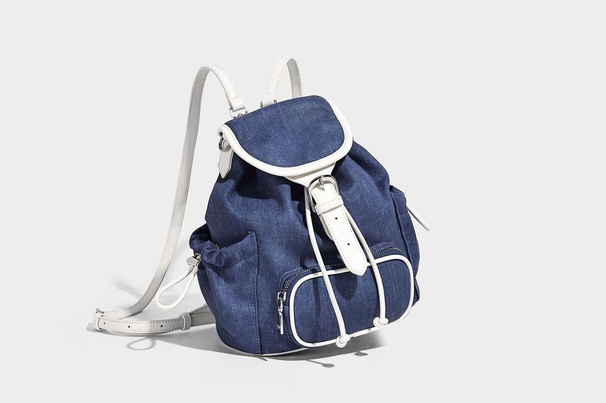 Compact denim drawstring backpack with white leather-look piping, flap-top buckle closure, front zip pocket, and silver-tone