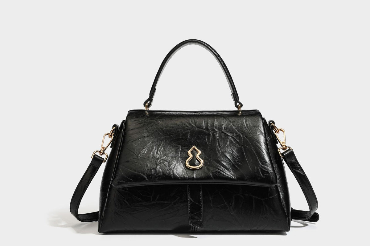 Structured black satchel bag with glossy crinkled leather-look finish, gold-tone emblem flap closure, slim top handle, and
