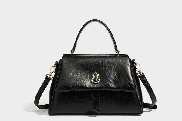 Structured black satchel bag with glossy crinkled leather-look finish, gold-tone emblem flap closure, slim top handle, and