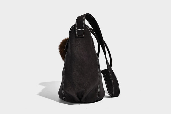 Small matte textured bucket crossbody bag with flap closure, gold-tone bar hardware, contrast stitching, and pompom tassel