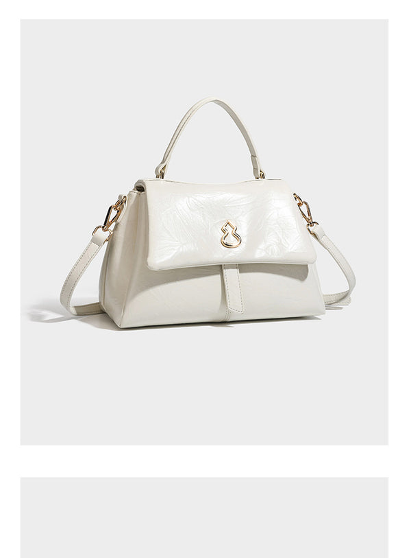 Structured black satchel bag with glossy crinkled leather-look finish, gold-tone emblem flap closure, slim top handle, and