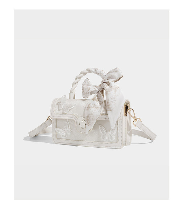 Small structured white flap bag with butterfly embroidery, flower-shaped turn-lock clasp, braided top handle with scarf bow
