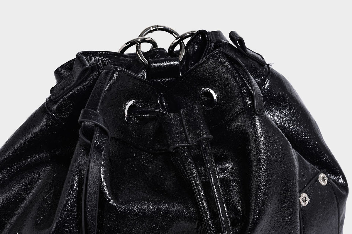 Glossy black convertible bucket backpack with silver grommet-studded straps, drawstring cinch closure, and front zip pocket