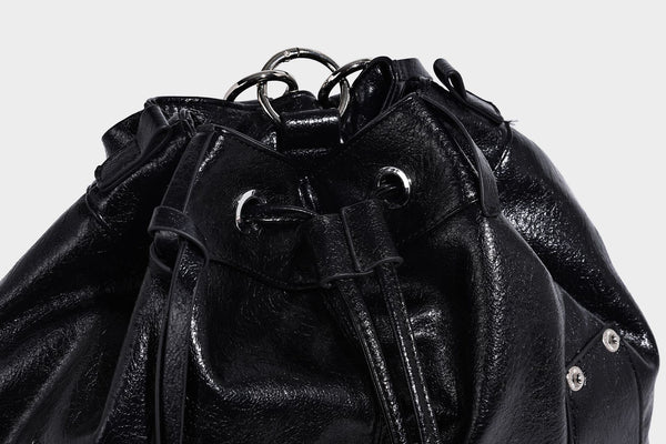 Glossy black convertible bucket backpack with silver grommet-studded straps, drawstring cinch closure, and front zip pocket