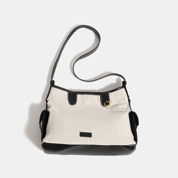 Off-white canvas shoulder bag with black leather-look trim, gold-tone ring hardware, top zip closure, side flap pocket, and