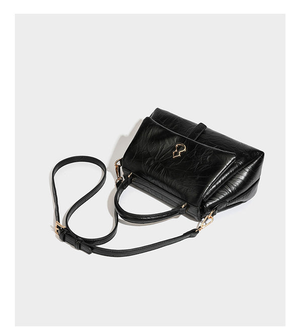 Structured black satchel bag with glossy crinkled leather-look finish, gold-tone emblem flap closure, slim top handle, and
