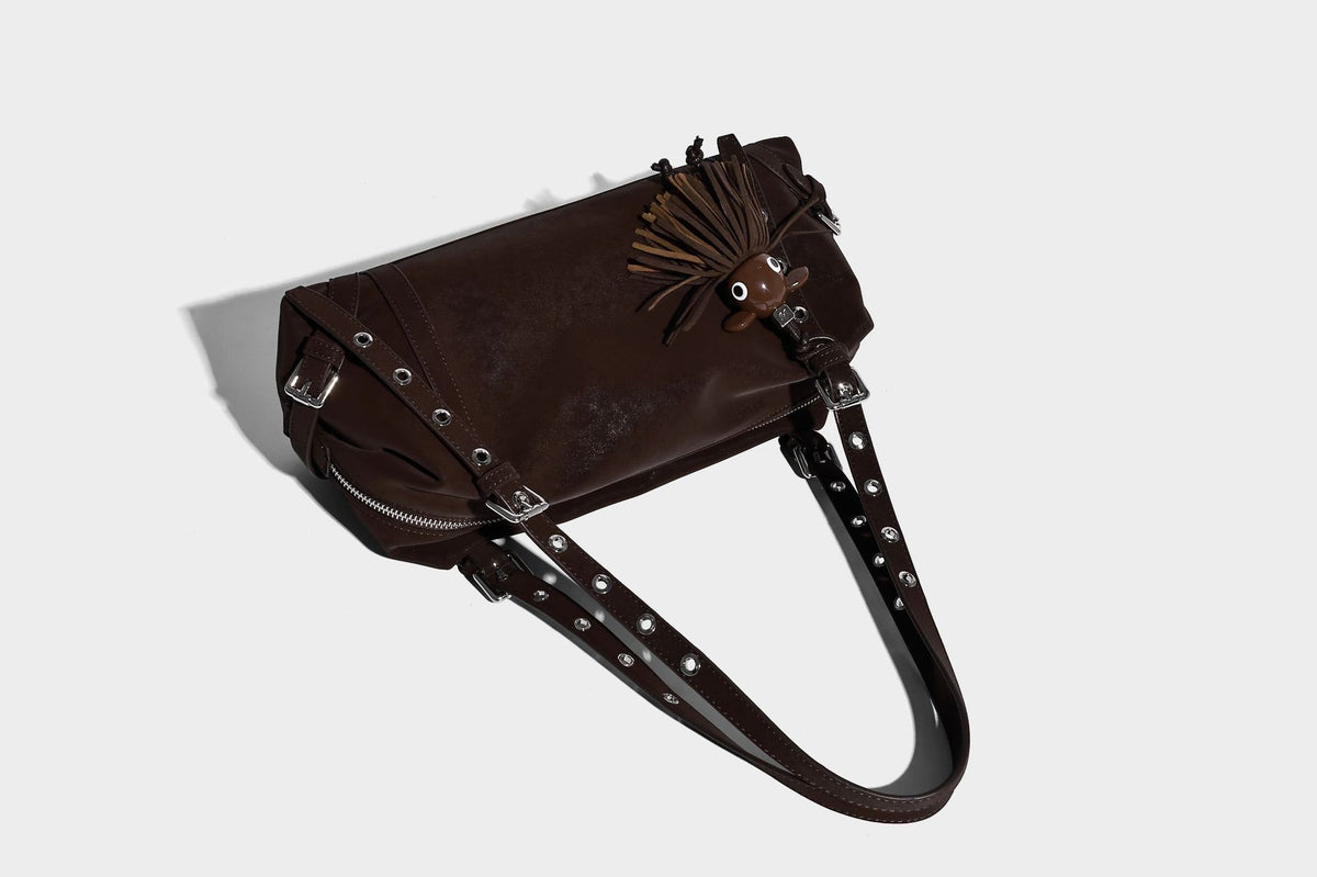 Weiche, zylinderförmige Schultertasche in Dunkelbraun mit silberfarbenen Riemen mit Ösen, Reißverschluss oben, seitlichen Schnallenbeschlägen und Quaste