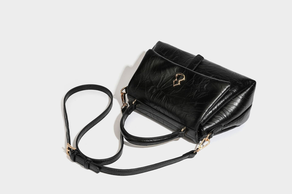 Structured black satchel bag with glossy crinkled leather-look finish, gold-tone emblem flap closure, slim top handle, and