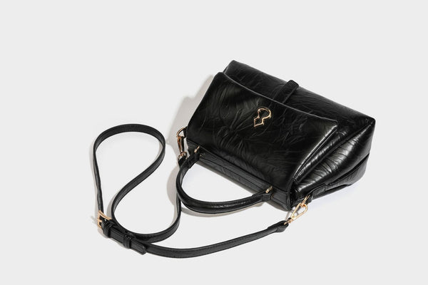 Structured black satchel bag with glossy crinkled leather-look finish, gold-tone emblem flap closure, slim top handle, and