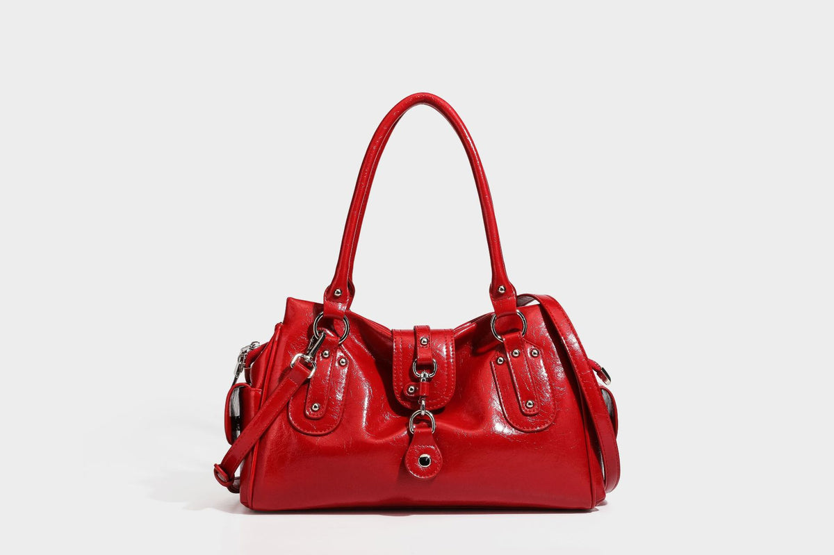 Glänzende, geknautschte Barrel-Satchel mit zwei oberen Tragegriffen, vorderem Klappen-Riemen-Panel, Ring- und Nietenbeschlägen sowie einem abnehmbaren