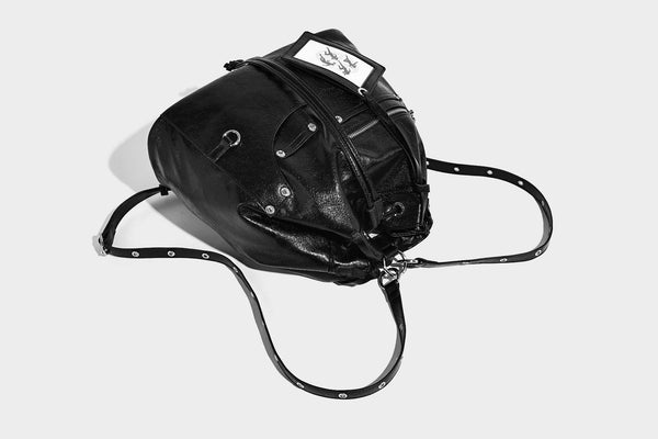 Glossy black convertible bucket backpack with silver grommet-studded straps, drawstring cinch closure, and front zip pocket