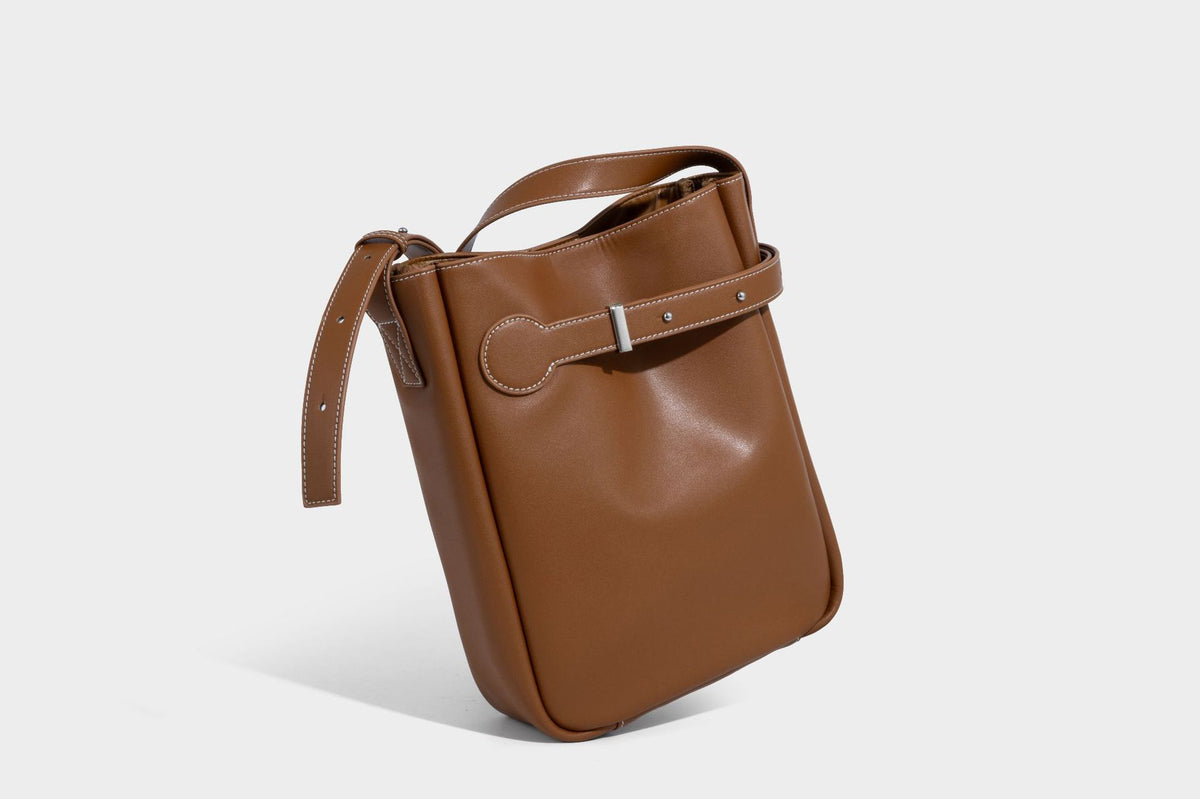 Structured dark brown leather-look bucket bag with white contrast stitching, front wraparound belt buckle strap, and long