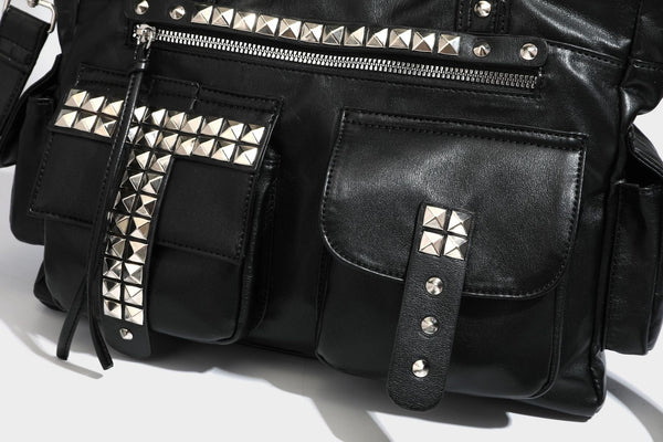 Black leather-look tote bag with silver pyramid studs on handles and front panel, multiple flap and zip exterior pockets