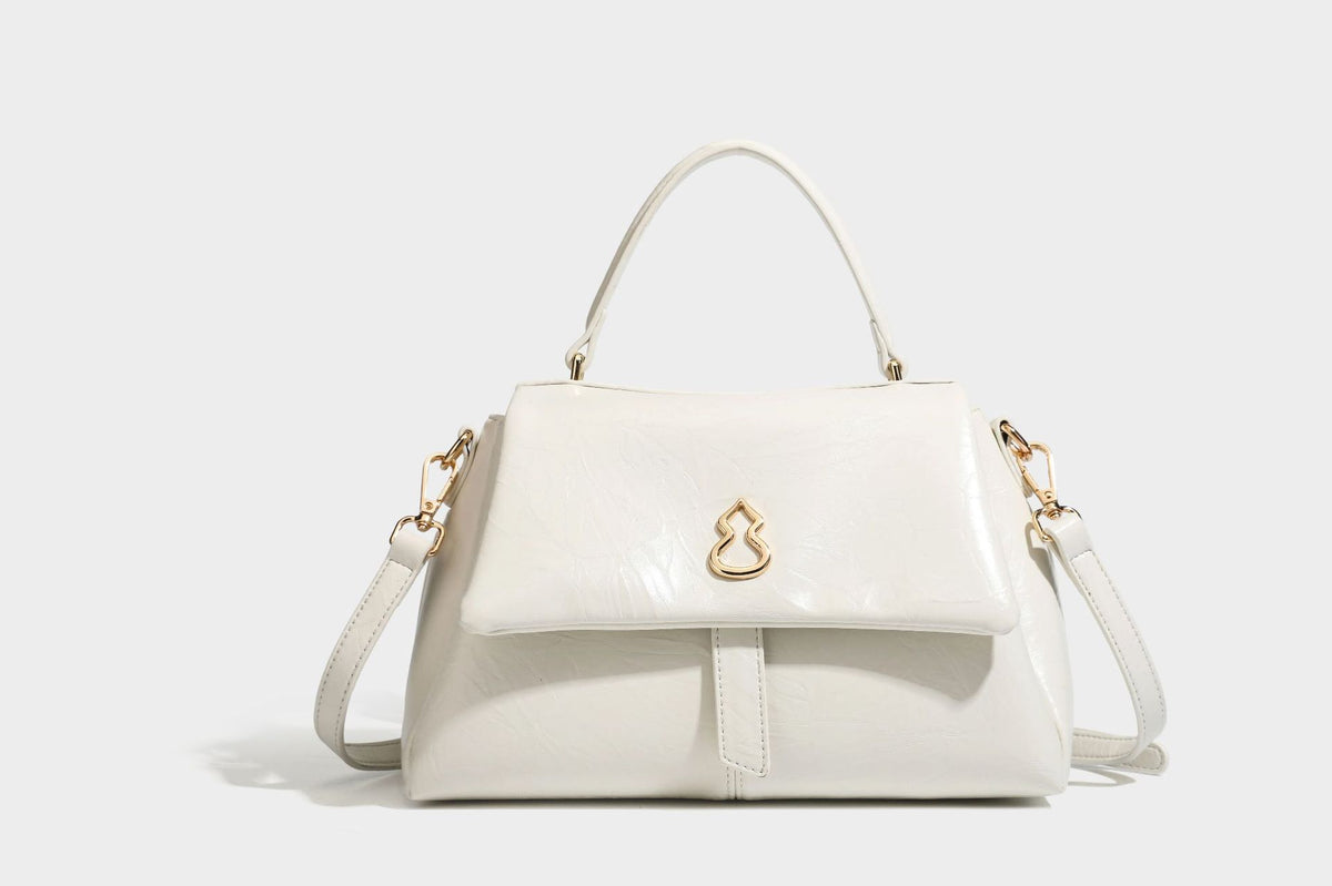 Structured black satchel bag with glossy crinkled leather-look finish, gold-tone emblem flap closure, slim top handle, and