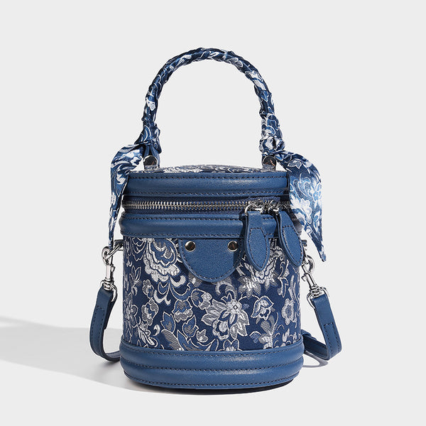 Small cylindrical barrel bag in blue-and-white floral jacquard with leather-look trim, scarf-wrapped top handle, silver-tone