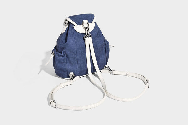 Compact denim drawstring backpack with white leather-look piping, flap-top buckle closure, front zip pocket, and silver-tone