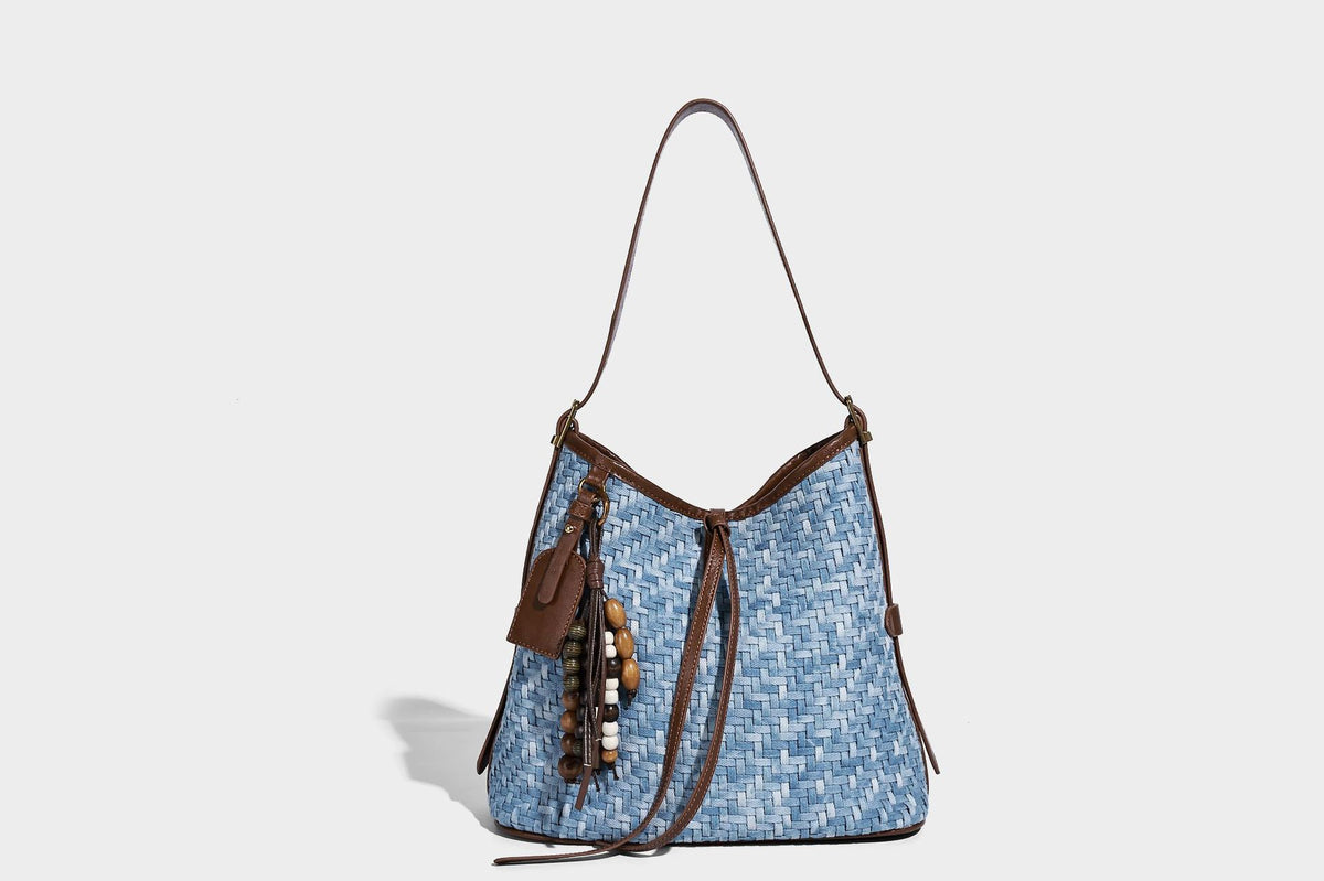 Light blue woven bucket bag with brown leather-look trim, gold-tone hardware, adjustable shoulder strap, and decorative bead