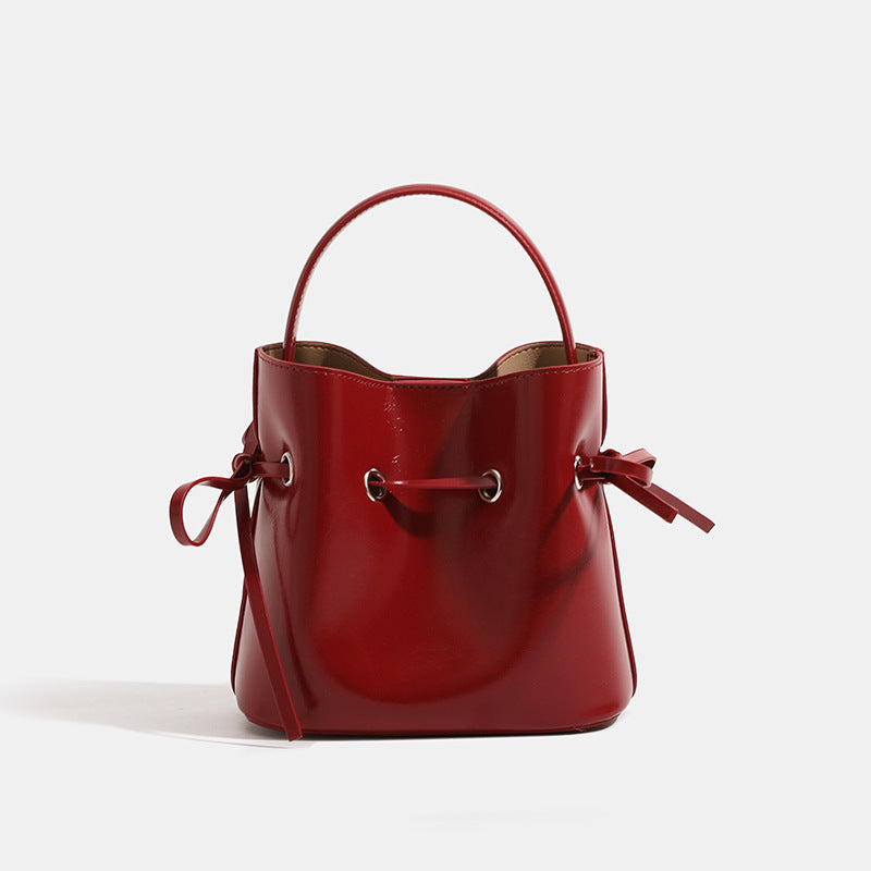 Compact glossy leather-look bucket bag with drawstring bow-tie closure, rounded top handle, and silver-tone grommet hardware