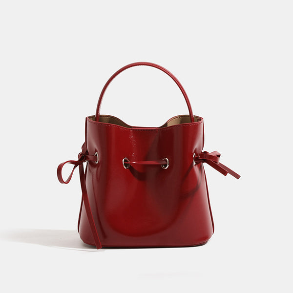 Compact glossy leather-look bucket bag with drawstring bow-tie closure, rounded top handle, and silver-tone grommet hardware