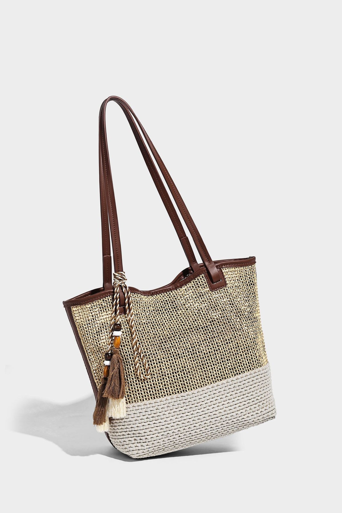 Woven beige and cream tote bag with brown leather-look trim, long double shoulder straps, and knotted tassel charm on front