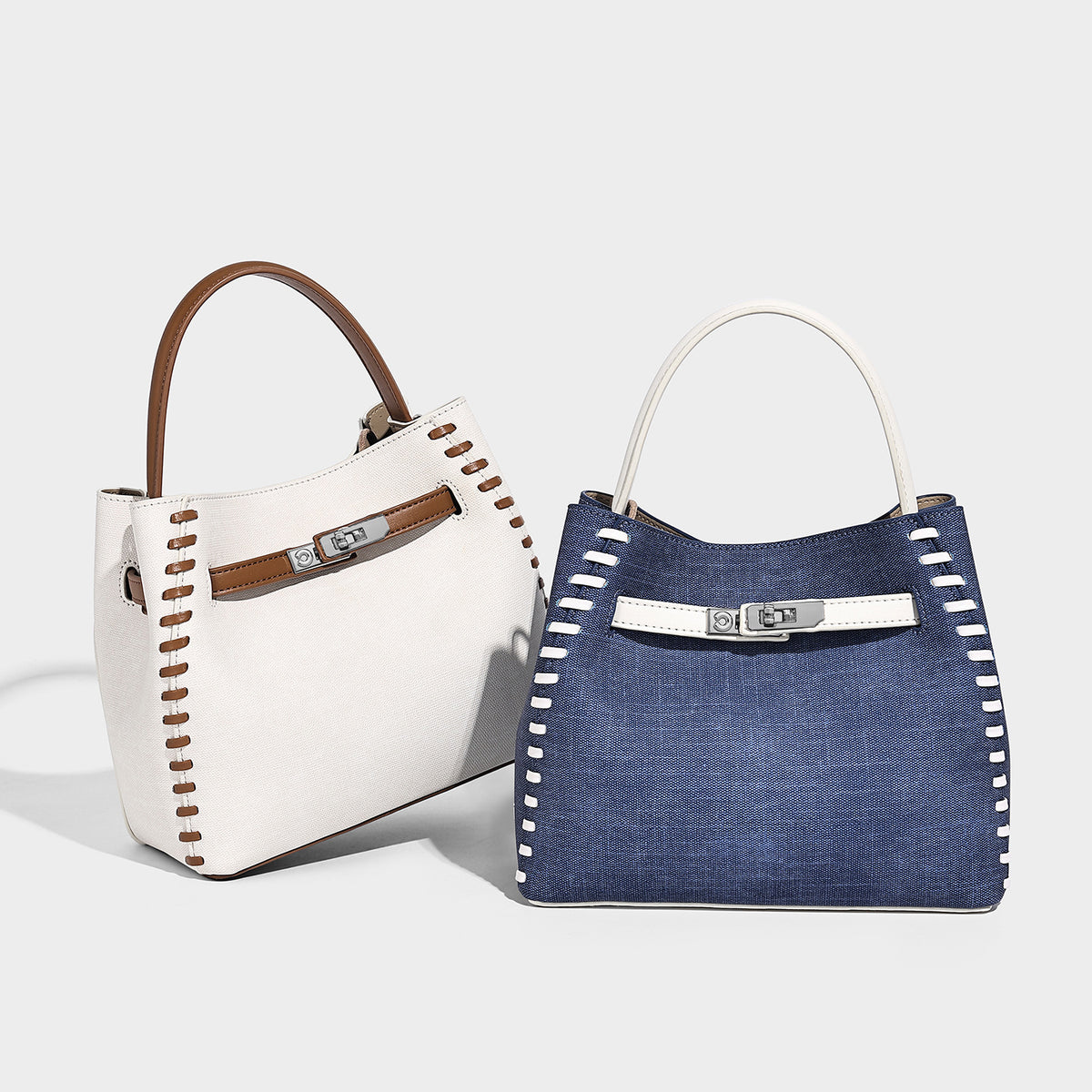 Structured denim bucket bag with white whipstitch trim, silver turn-lock hardware, short top handles, and a long adjustable