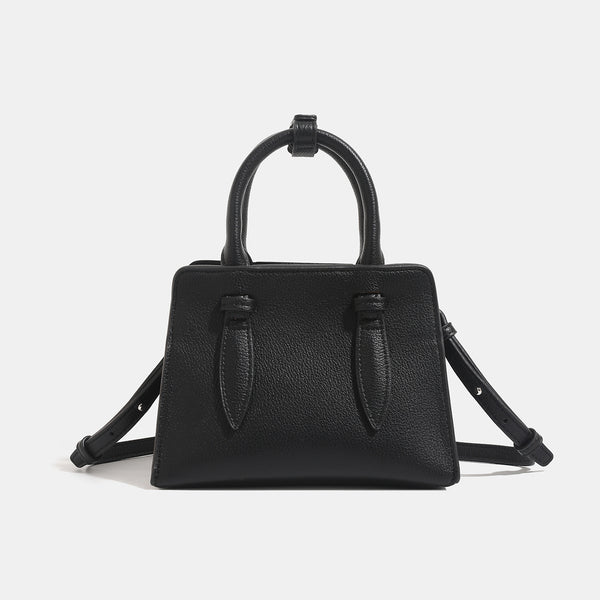 Small structured trapezoid top-handle bag in pebbled matte finish with decorative strap tabs, twin knotted handles, and