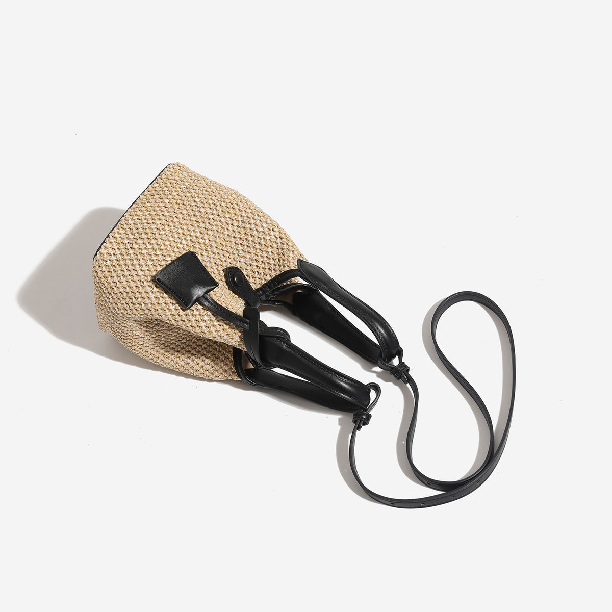 Woven beige straw bucket bag with black leather-look trim, dual top handles, detachable shoulder strap, and front snap