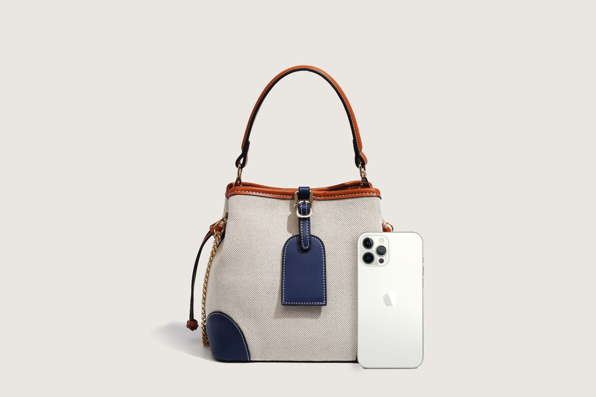 Cream woven-texture canvas bucket bag with navy accent panels, tan leather-look trim, gold-tone chain strap, and tassel