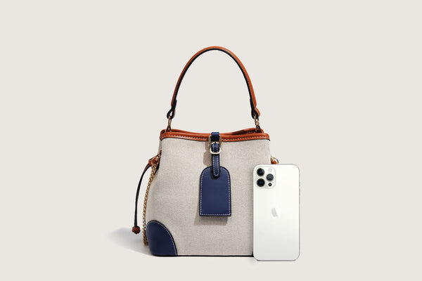 Cream woven-texture canvas bucket bag with navy accent panels, tan leather-look trim, gold-tone chain strap, and tassel