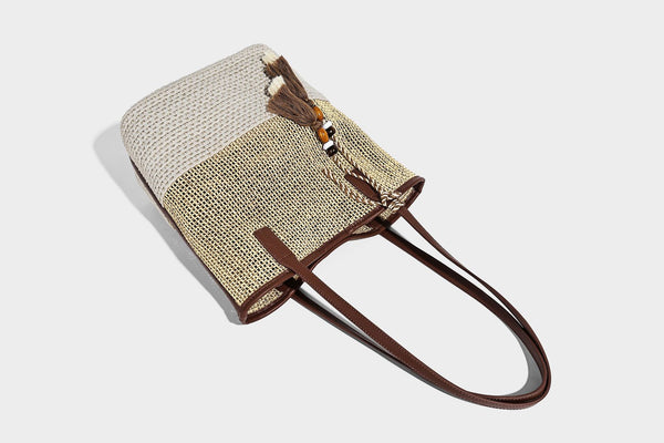Woven straw-look tote bag in tan and cream with dark brown leather-look trim, long flat shoulder straps, and tassel bead