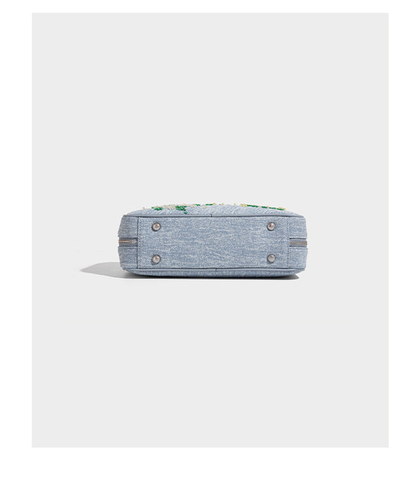 Small structured light blue denim-texture bowling bag with white floral bead embroidery, dual rounded top handles, and