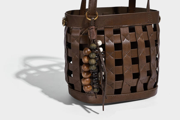 Small woven lattice bucket bag in brown leather-look finish with beaded tassel charm, dual top handles, and detachable