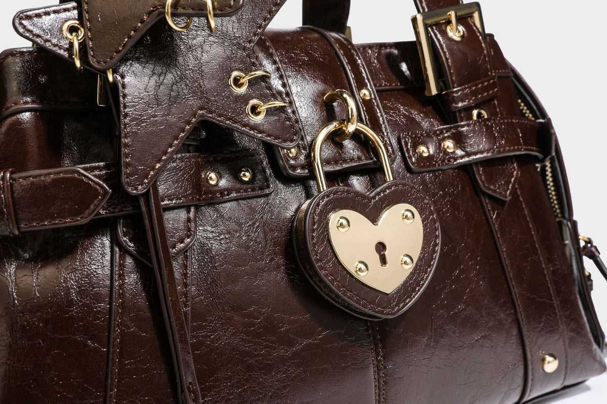 Structured leather-look satchel bag with heart padlock charm, star charm, gold-tone buckle hardware, dual top handles, and