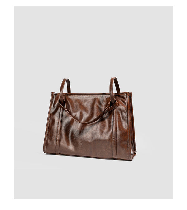 Large structured glossy leather-look tote bag with dual top handles and long shoulder strap in burnished brown, detail view