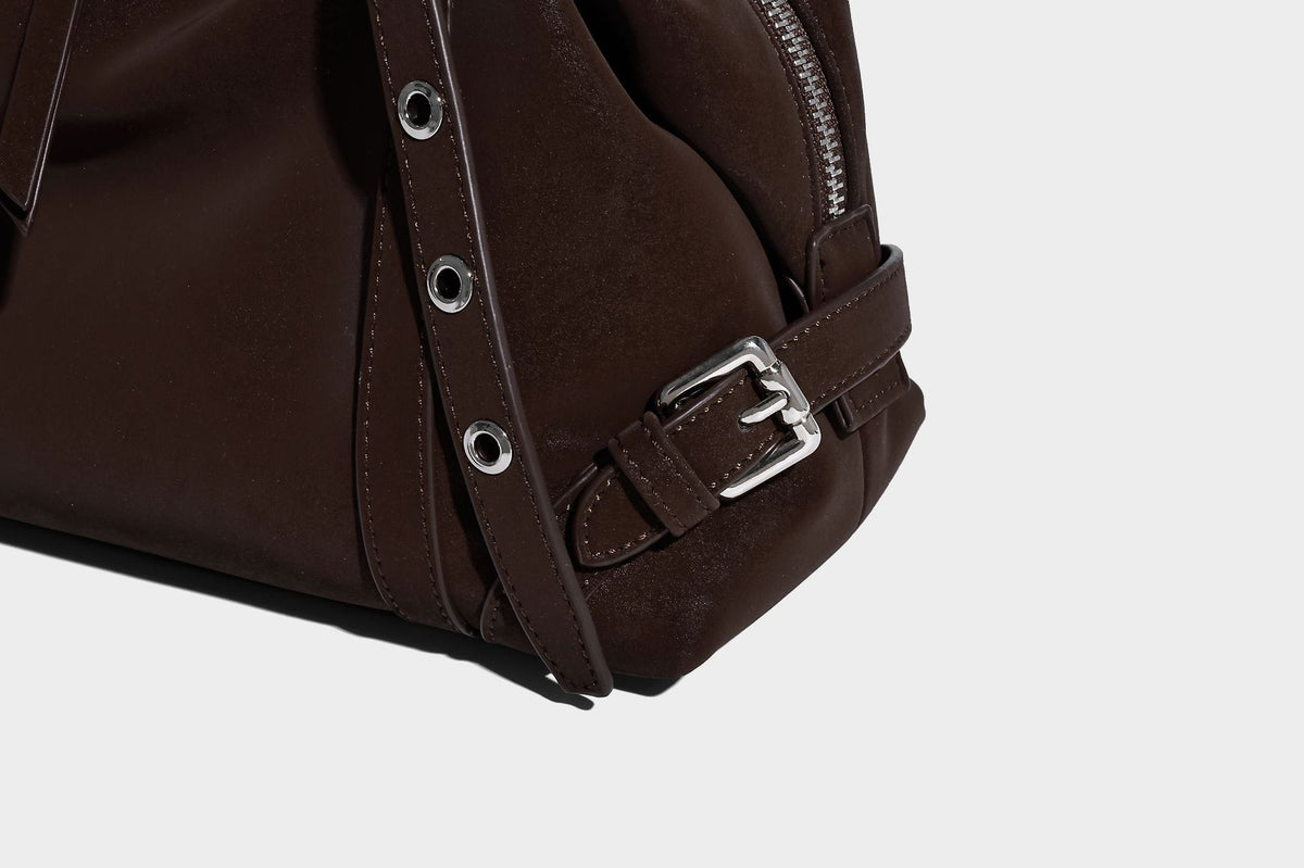 Weiche, zylinderförmige Schultertasche in Dunkelbraun mit silberfarbenen Riemen mit Ösen, Reißverschluss oben, seitlichen Schnallenbeschlägen und Quaste