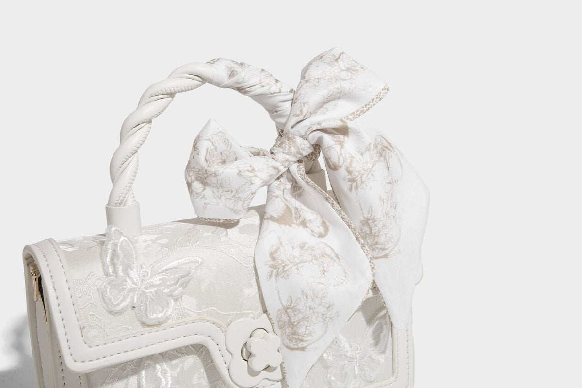 Small structured white flap bag with butterfly embroidery, flower-shaped turn-lock clasp, braided top handle with scarf bow
