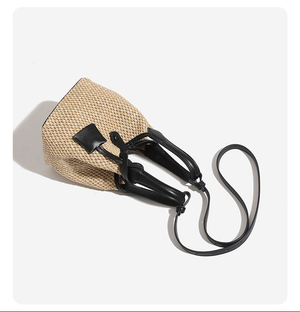 Woven beige straw bucket bag with black leather-look trim, dual top handles, detachable shoulder strap, and front snap