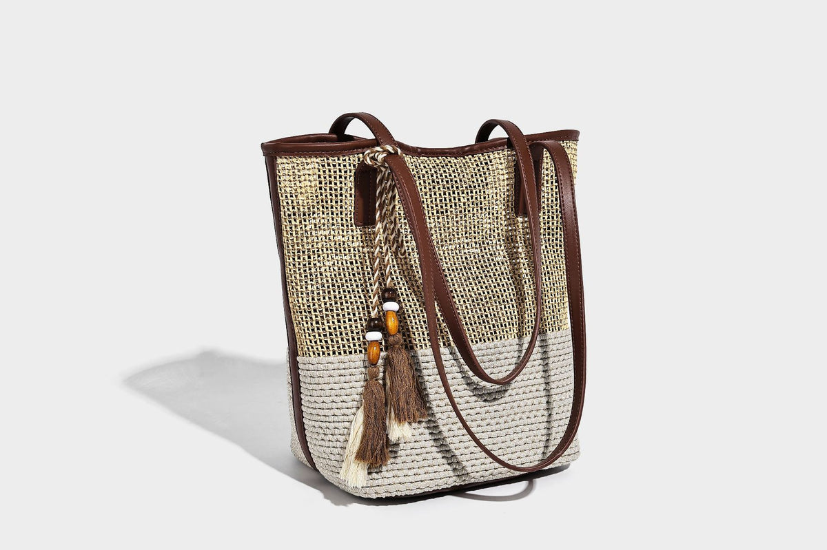 Woven straw-look tote bag in tan and cream with dark brown leather-look trim, long flat shoulder straps, and tassel bead