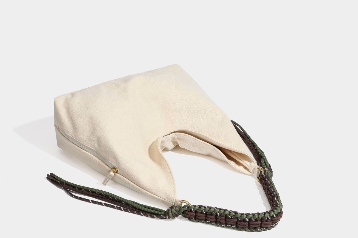 Soft slouchy cream canvas shoulder tote with braided brown and green cord handle, gold-tone ring hardware, and tassel trim