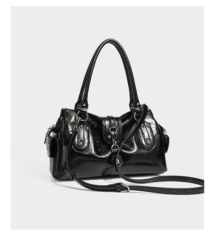 Glänzende, geknautschte Barrel-Satchel mit zwei oberen Tragegriffen, vorderem Klappen-Riemen-Panel, Ring- und Nietenbeschlägen sowie einem abnehmbaren
