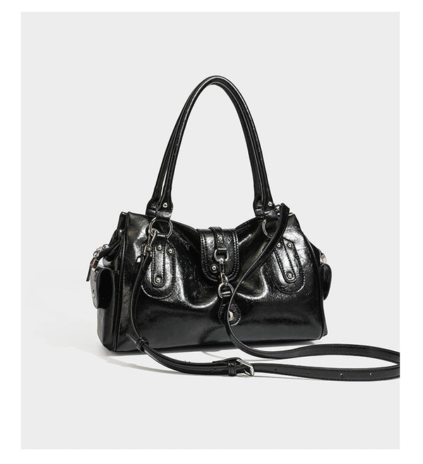 Glänzende, geknautschte Barrel-Satchel mit zwei oberen Tragegriffen, vorderem Klappen-Riemen-Panel, Ring- und Nietenbeschlägen sowie einem abnehmbaren