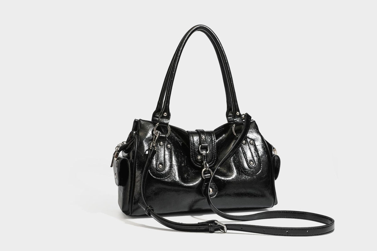 Glänzende, geknautschte Barrel-Satchel mit zwei oberen Tragegriffen, vorderem Klappen-Riemen-Panel, Ring- und Nietenbeschlägen sowie einem abnehmbaren
