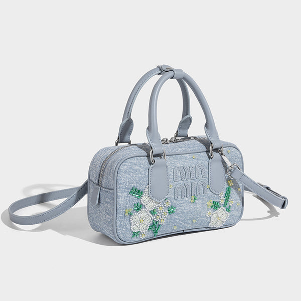 Small light blue denim-look barrel bag with white floral bead embroidery, dual top handles, and detachable shoulder strap