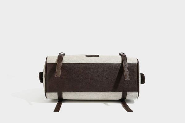 Beige canvas barrel bag with dark brown leather-look trim, silver-tone hardware, top zip closure, and adjustable shoulder