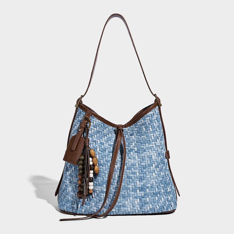 Light blue woven bucket bag with brown leather-look trim, gold-tone hardware, adjustable shoulder strap, and decorative bead