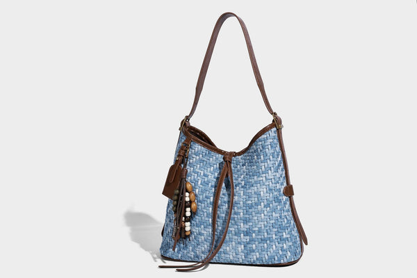 Light blue woven bucket bag with brown leather-look trim, gold-tone hardware, adjustable shoulder strap, and decorative bead