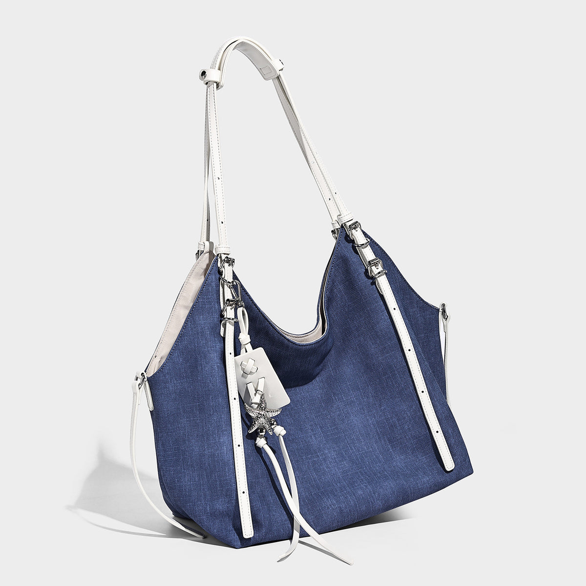 Large slouchy denim-look canvas shoulder tote bag with white leather-look straps, silver-tone buckle hardware, and hanging