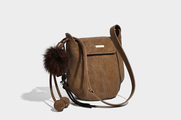 Small matte textured bucket crossbody bag with flap closure, gold-tone bar hardware, contrast stitching, and pompom tassel