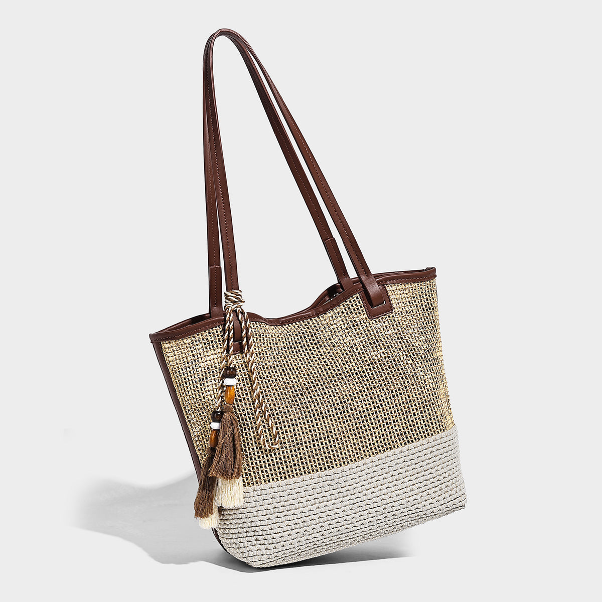 Woven beige and cream tote bag with brown leather-look trim, long double shoulder straps, and knotted tassel charm on front
